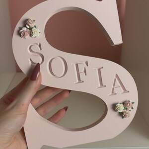 May include: A pale pink letter S with the name SOFIA engraved in capital letters across the centre. The letter is decorated with small clusters of artificial cream and dusty pink roses positioned at the top left and bottom right corners.