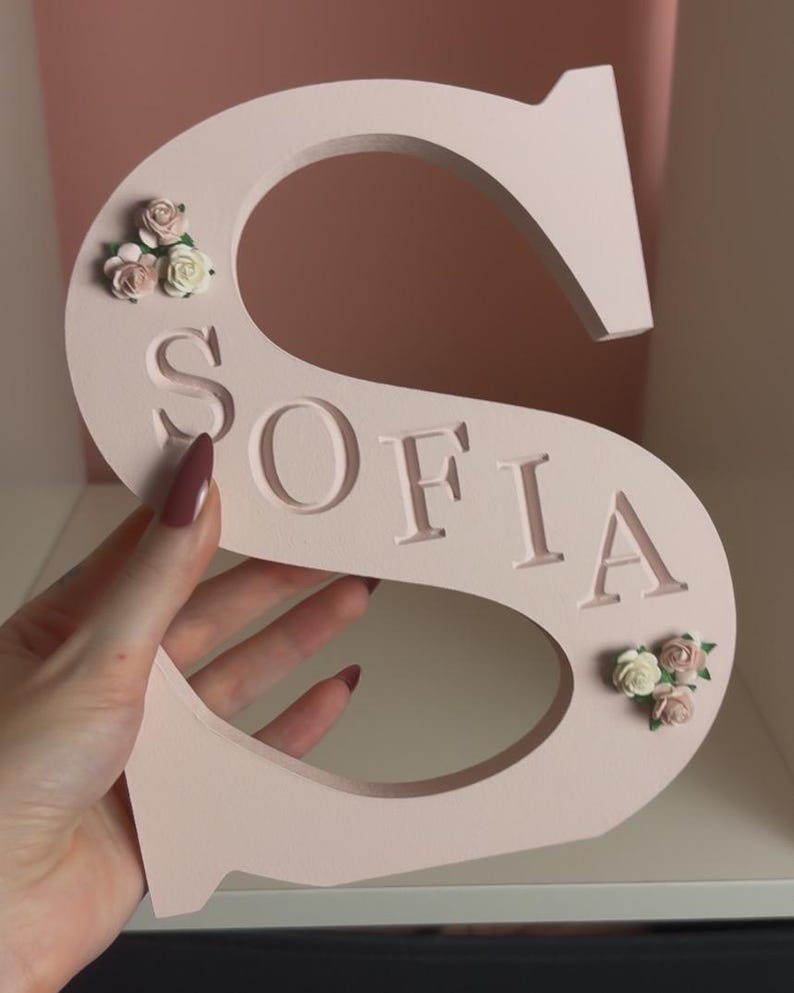 May include: A pale pink letter S with the name SOFIA engraved in capital letters across the centre. The letter is decorated with small clusters of artificial cream and dusty pink roses positioned at the top left and bottom right corners.