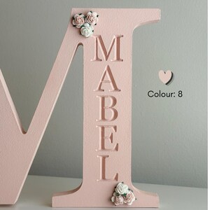 May include: A large, light pink wooden letter M featuring the name MABEL engraved vertically in a serif font. The letter is decorated with small, artificial white and pink roses at the top left and bottom right corners, with the text Colour: 8.