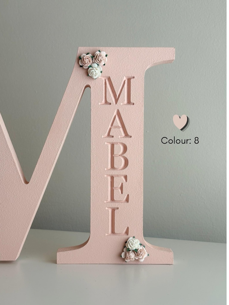 May include: A large, light pink wooden letter M featuring the name MABEL engraved vertically in a serif font. The letter is decorated with small, artificial white and pink roses at the top left and bottom right corners, with the text Colour: 8.