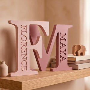 May include: Two large, freestanding wooden letters, an F and an M, finished in shades of pink. The letter F features the name FLORENCE engraved vertically, while the letter M features the name MAYA engraved vertically in a clean, sans-serif font.