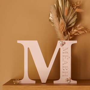 May include: A light pink wooden letter M featuring the name M&Eacute;ABH engraved vertically on the right side. The letter is decorated with small clusters of miniature cream and pink fabric flowers at the top and bottom of the right vertical bar.
