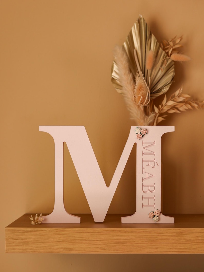 May include: A light pink wooden letter M featuring the name M&Eacute;ABH engraved vertically on the right side. The letter is decorated with small clusters of miniature cream and pink fabric flowers at the top and bottom of the right vertical bar.