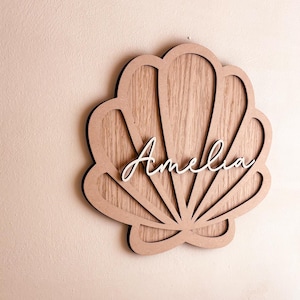 May include: A decorative wall sign shaped like a seashell, crafted from light brown wood with a visible grain. The centre of the shell features the name Amelia in white, raised, cursive script lettering, creating a layered, dimensional appearance.