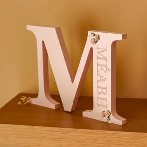 May include: A light pink, freestanding letter M featuring the name M&Eacute;ABH engraved vertically along the right side. The letter is decorated with small, artificial cream and peach-coloured roses positioned at the top and bottom of the right stem.