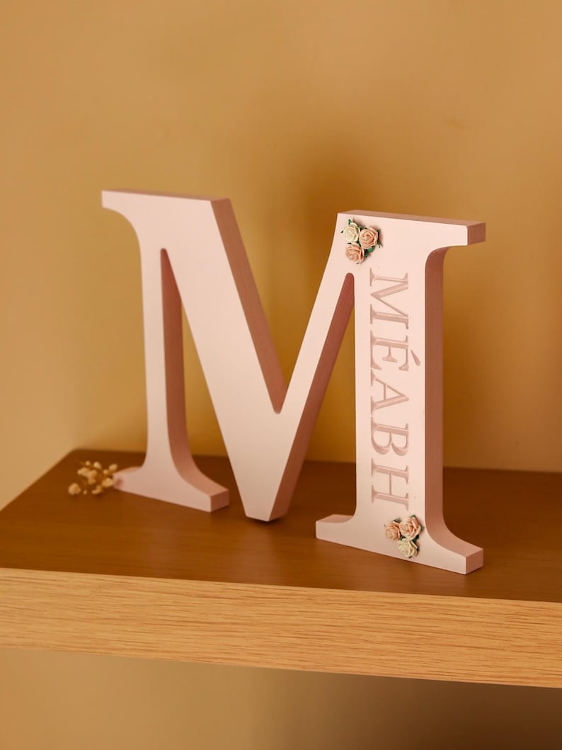 May include: A light pink, freestanding letter M featuring the name M&Eacute;ABH engraved vertically along the right side. The letter is decorated with small, artificial cream and peach-coloured roses positioned at the top and bottom of the right stem.