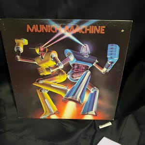 Könnte beinhalten: Ein Vinyl-Plattencover mit zwei tanzenden Robotern. Die Roboter sind blau und gelb mit leuchtend roten Augen. Das Cover ist schwarz mit dem Text "MUNICH MACHINE" in roten Buchstaben.