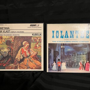May include: Two vintage vinyl record albums. The album on the left is labeled "Smetana Ma Vlast Complete Recording" with artwork of figures in traditional dress. The album on the right is labeled "Iolanthe" with a stage scene featuring costumed figures.