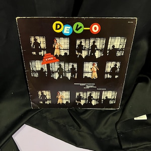 May include: A black vinyl record album cover with the band name "DEVO" in colorful letters. The cover features a series of black and white photos of the band members performing on stage. The album title "LIVE" is printed in red on a white background.