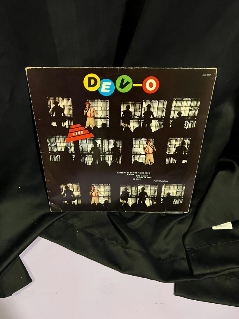 Pu&ograve; includere: Una copertina di album in vinile nero con il nome della band "DEVO" in lettere colorate. La copertina presenta una serie di foto in bianco e nero dei membri della band che si esibiscono sul palco. Il titolo dell'album "LIVE" &egrave; stampato in rosso su sfondo bianco.