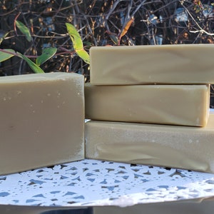 May include: Four bars of handmade soap stacked on a silver tray with a white doily. The soap is a light beige color.