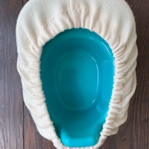 May include: A light blue plastic bedpan covered with a cream-colored, fitted, cloth cover. The bedpan is oval-shaped and sits on a dark brown wooden surface. The cloth cover is gathered around the rim of the bedpan.
