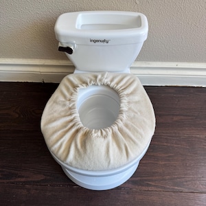 May include: A white plastic Ingenuity potty training toilet featuring a beige fabric seat cover with an elasticized edge. The toilet has a black flush handle on the side and a removable inner basin, designed for children learning to use the restroom.