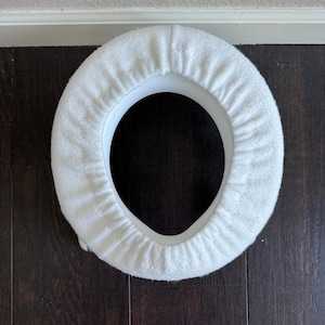 May include: A white, plush toilet seat cover with a soft, textured surface. The cover is circular with a white plastic base and is designed to fit a standard toilet seat. The cover is placed on a dark wood floor.