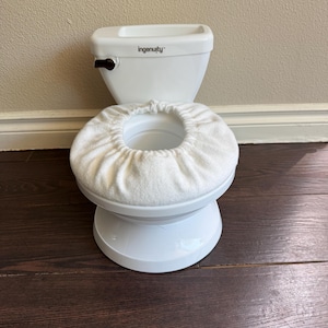 May include: A white plastic potty training toilet with a dark brown flush handle and the brand name Ingenuity printed on the tank. The seat is covered with a white fabric cover that has an elasticized opening in the center.
