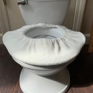 May include: A white Summer brand toilet with a white plush toilet seat cover. The toilet is set against a neutral wall and dark wood floor. The toilet has a chrome flush handle.