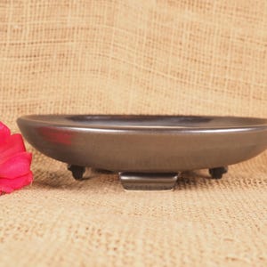 May include: A dark brown ceramic bonsai pot with a low, wide design. The pot has a rounded shape and sits on three small feet. A bright pink flower is visible on the left side of the image.