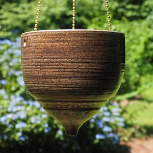 May include: A brown ceramic hanging planter with a tapered, pointed bottom and horizontal ridged texture. The vessel features three small holes near the top rim for suspension and is attached to thin gold-toned metal ball chains for hanging.