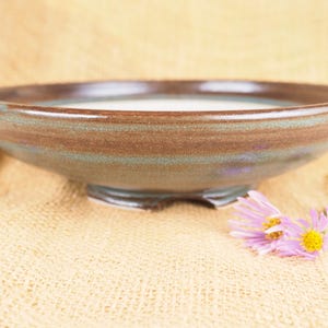 May include: A shallow, round ceramic bowl with a wide rim, featuring a smooth, glossy glaze in earthy tones of brown and muted green. The bowl has a raised footed base and a light-colored interior, showcasing a simple, functional design.