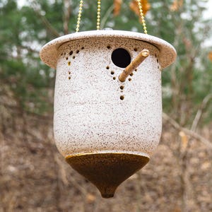 May include: A speckled ceramic birdhouse with a brown base and a small wooden perch. The birdhouse is suspended by a gold chain and has a round opening for birds to enter. The exterior is a speckled white and brown.