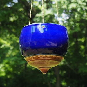 May include: A hanging ceramic planter with a glossy, deep cobalt blue upper section and a contrasting unglazed, textured tan base that tapers to a point. The planter is suspended by thin gold-toned metal chains attached to the rim.