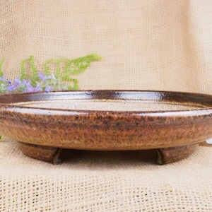 May include: A shallow, round ceramic dish with a speckled, light-colored interior and a dark, mottled brown exterior glaze. The dish features a wide, slightly curved rim and is supported by three short, sturdy feet integrated into the base.