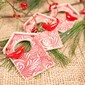 May include: Four red and white ceramic birdhouse ornaments with red birds inside. The ornaments are on a burlap background with pine needles.