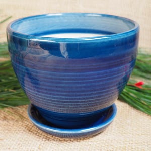 May include: A blue ceramic planter with a wide, round base and a saucer. The planter has a glazed finish with a subtle striped pattern.