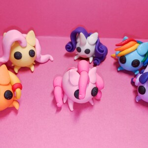 May include: A collection of six colorful, handcrafted clay figurines resembling cartoon ponies. Each figure has a unique color scheme, including yellow, pink, blue, purple, and orange, with detailed manes and tails. The figurines are set against a pink background.