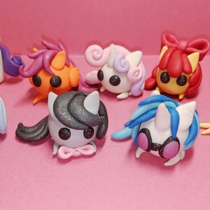 May include: A collection of eight colorful, handcrafted clay figurines of stylized ponies. Each pony has a unique color scheme, including white, blue, orange, pink, and gray, with matching manes and tails. The ponies have large, black eyes and are set against a pink background.