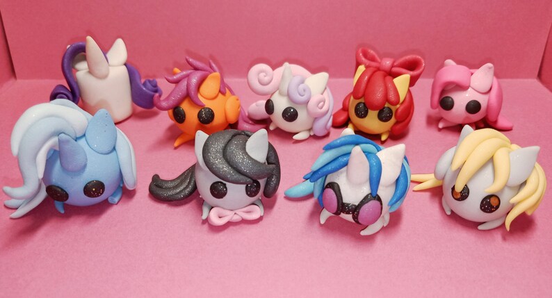 May include: A collection of eight colorful, handcrafted clay figurines of stylized ponies. Each pony has a unique color scheme, including white, blue, orange, pink, and gray, with matching manes and tails. The ponies have large, black eyes and are set against a pink background.