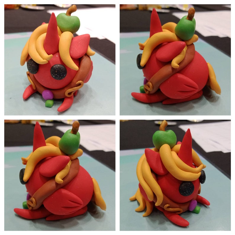 May include: A whimsical, handcrafted clay unicorn figurine. The unicorn is primarily red with yellow, brown, green, and purple accents. It features a horn, flowing mane, and a small green apple on its head. The eyes are black with glitter.