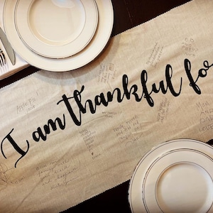 May include: A beige table runner with the phrase "I am thankful for..." in large black script. Handwritten notes and drawings surround the text. Two white plates with gold rims and silverware are on the table.