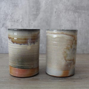May include: Two cylindrical ceramic tumblers. The tumblers have a neutral color palette with shades of beige, brown, and orange. The rims are dark, and the bases are a reddish-orange. The tumblers are handmade and have a rustic appearance.