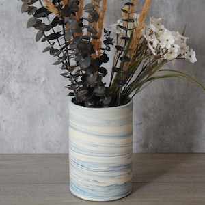May include: A cylindrical ceramic vase featuring a marbled pattern of white and soft blue swirls. The vase is filled with a decorative arrangement of dried dark eucalyptus branches, tall golden wheat stalks, and small white flowers.