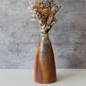 May include: A ceramic vase with a narrow neck and a wide base, featuring a gradient of brown and orange hues. The vase holds a bouquet of dried flowers and grasses, creating a rustic and natural aesthetic.