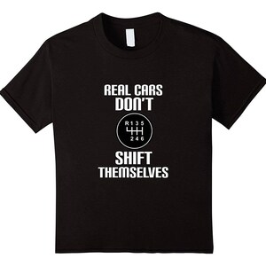 May include: Black t-shirt with white text that reads "Real Cars Don't Shift Themselves". A white gear shift pattern is in the center of the shirt.