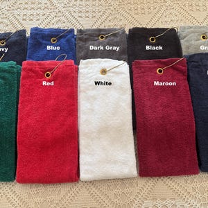 May include: A selection of golf towels in a range of colours: navy, blue, dark grey, black, grey, green, red, white, maroon, and navy. Each towel has a gold-coloured grommet and a loop for hanging.