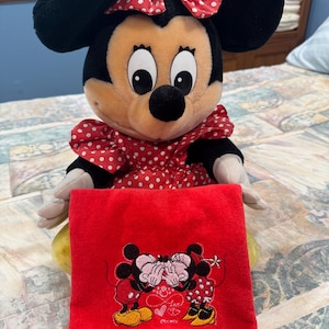 Puede incluir: Una muñeca de peluche de Minnie Mouse con orejas negras, cara bronceada y un vestido rojo con lunares blancos. La muñeca sostiene una pieza de tela rectangular roja con un bordado de dos ratones besándose y el texto Nathan & Anna debajo.