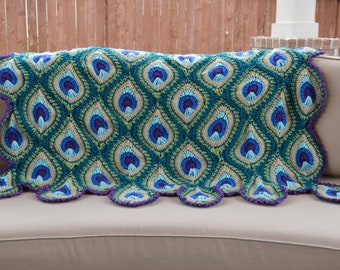 May include: A crocheted peacock feather throw blanket in shades of teal, green, blue, and purple. The blanket has a scalloped edge and is draped over a light brown couch.