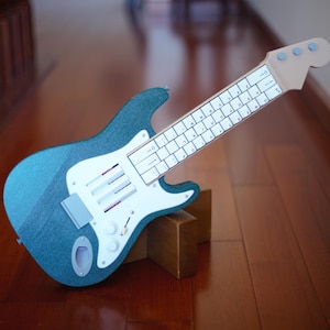 Op de afbeelding: Een blauwgroen glitterend object in de vorm van een elektrische gitaar met een witte slagplaat met drie verticale sleuven en een toets ontworpen als een computertoetsenbord met witte toetsen en zwarte letters, inclusief drie lichtblauwe stemmechanieken.