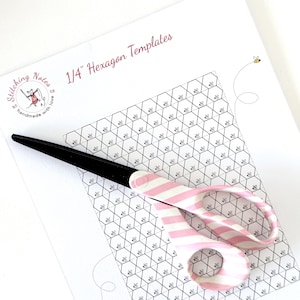 May include: A white paper sheet titled 1/4" Hexagon Templates featuring a black and white grid of hexagons with small bee illustrations, and a pair of scissors with black blades and a pink and white striped handle resting on top of the paper.