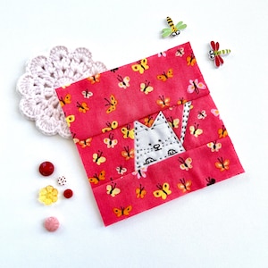 May include: A pink fabric square with a white cat appliqué. The cat is surrounded by small yellow butterflies. The fabric is decorated with a repeating pattern of butterflies.