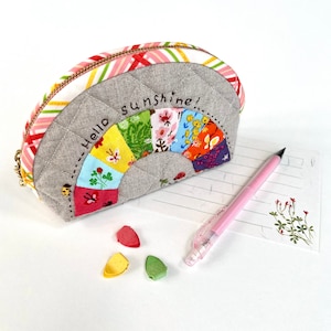 Może przedstawiać: Pikowana, półokrągła kosmetyczka z tęczowym wzorem i napisem "Hello sunshine!" wyhaftowanym z przodu. Kosmetyczka ma złoty zamek błyskawiczny i wzorzyste wykończenie z tkaniny. Obecny jest również różowy ołówek i trzy kolorowe klipsy.