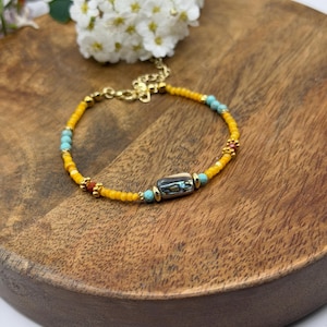 Puede incluir: Una pulsera de cuentas con detalles amarillos, turquesas y dorados. La pulsera presenta una cuenta rectangular central con colores iridiscentes. La pulsera se exhibe sobre una superficie de madera, con flores blancas en el fondo.
