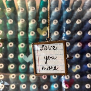 May include: A small, square, framed piece with the embroidered words "Love you more" on lined paper. The frame is suspended by a thin, brown string. The background features a gradient of spools of thread in shades of blue and green.