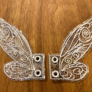 May include: A pair of clear, sparkly fairy wings with white embroidery. The wings have eyelets on the sides for attaching to a costume.