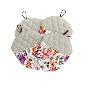 Peut inclure: Ensemble de quatre maniques matelassées en forme de glands. Les maniques sont en tissu de lin beige avec un imprimé floral sur le devant. Le dos des maniques est de couleur beige unie.