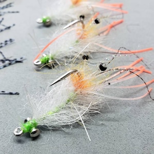 May include: A close-up of a white and orange shrimp fly fishing lure with black bead eyes and a green bead head. The lure has a long, thin body and several long, thin legs.