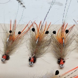 May include: Four brown and orange shrimp fly fishing lures with black beads for eyes. The lures have a brown and white feather body and orange legs.